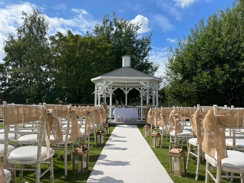 An outdoor ceremony on the lawn, a white gazebo at the end of an aisle runner lined with hessian-tied chiavari chairs
