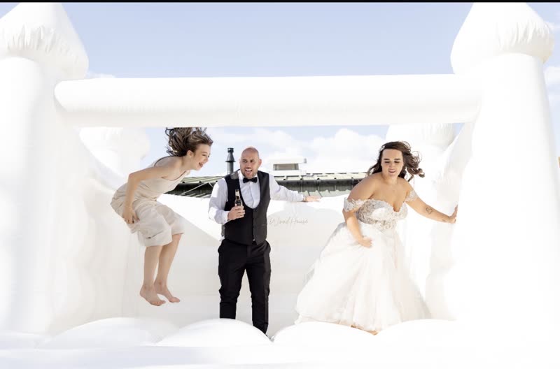 A white inflatable bouncy castle on the grounds with a wedding party mid-jump inside it