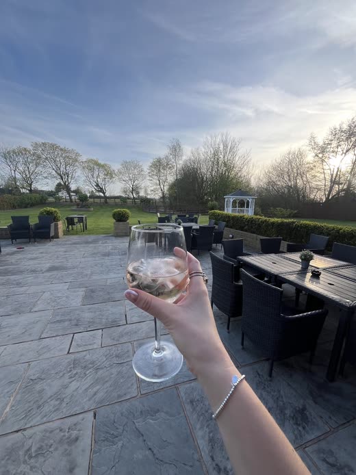 A hand holding a cocktail up over the venue's paved terrace at dusk, with rattan furniture and open fields beyond